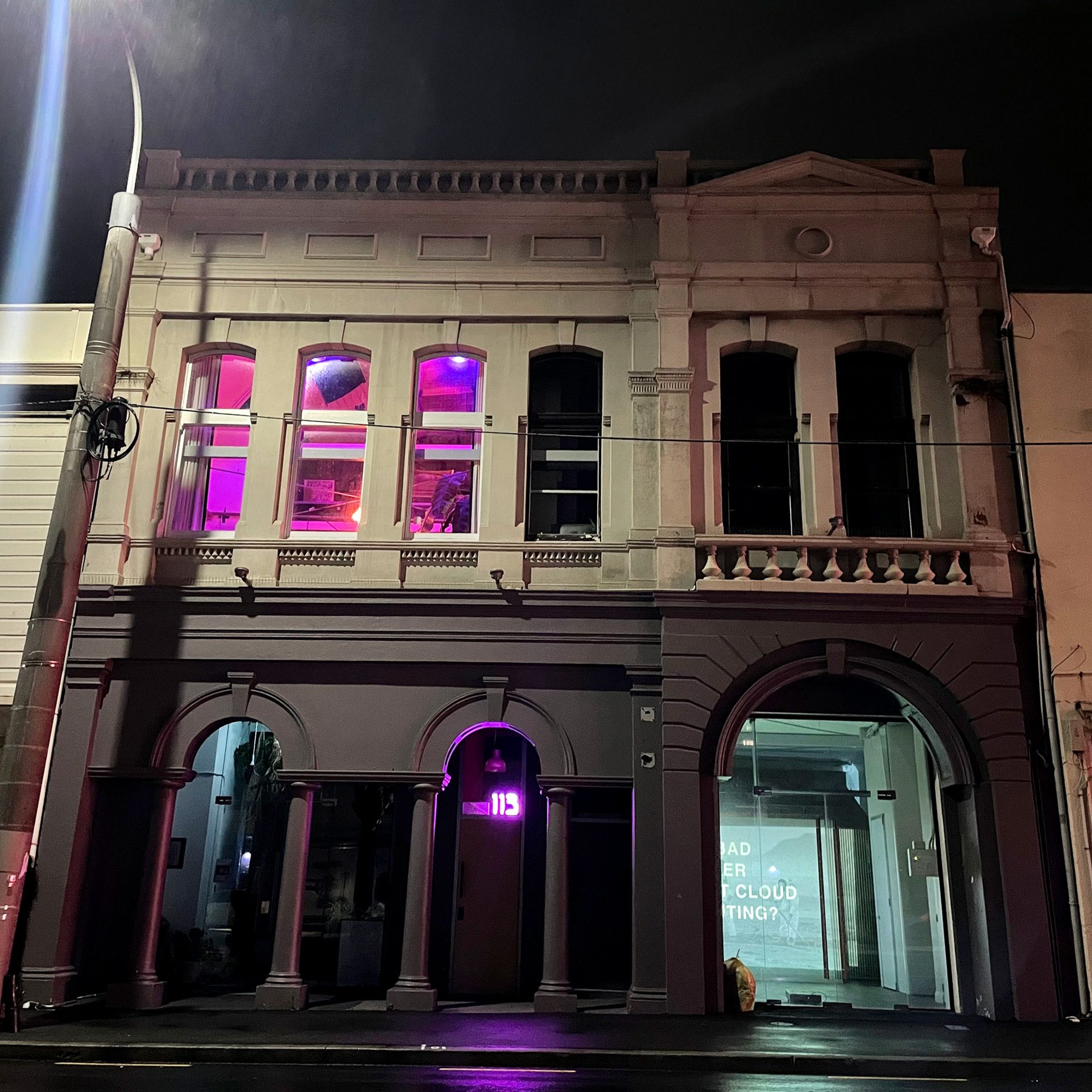 The exterior of the building, a two-story stone building with arched doors and many windows. It is night-time, and pink lighting spills out of an upstairs room.